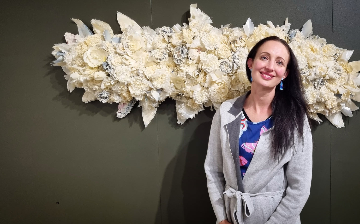 Vanessa stands smiling in front of her cream wall installation artwork on grey wall.